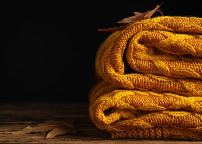 Orange knitted plaid with dry leaves on wooden table, closeup. Space for text